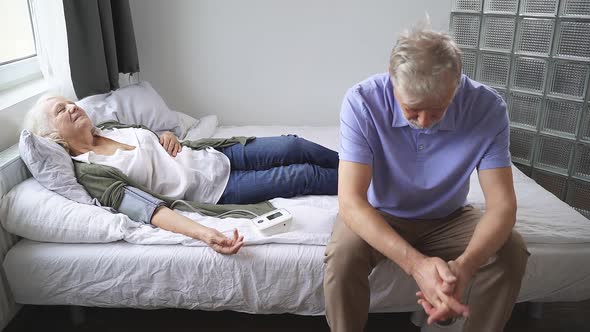 Woman of Retirement Age Is Lying on a Bed Due To High Blood Pressure and Her Husband Is Sitting Next