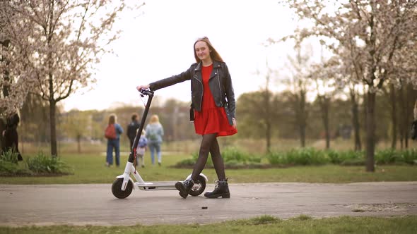 Young Stylish Woman Stands and Holds Electric Scooter in City Park