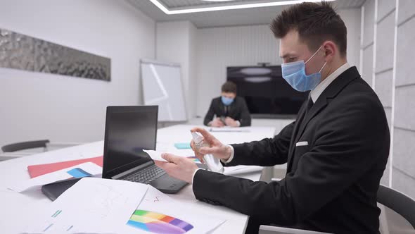 Side View of Serious Caucasian Employee Disinfecting Laptop with Sanitizer Sitting in Office with