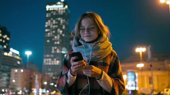 Lady with Earphones Walks Listening To Music on Phone in City with Night Lights