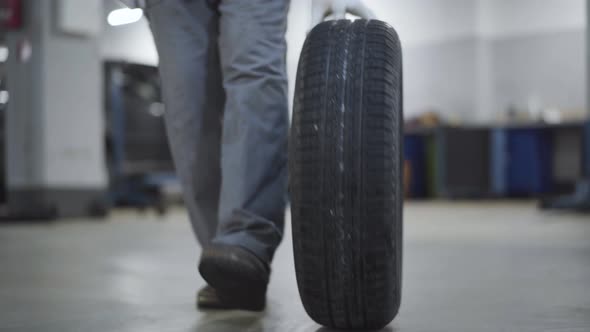 Unrecognizable Worker Rolling Car Wheel in Repair Shop. Man in Workwear and Protective Gloves