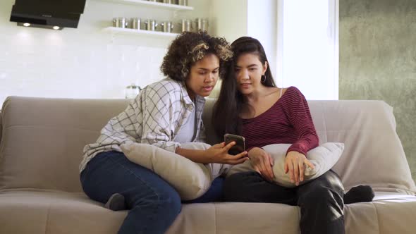 Happy Girlfriends Looking At Smartphone Sitting On Couch At Home Together