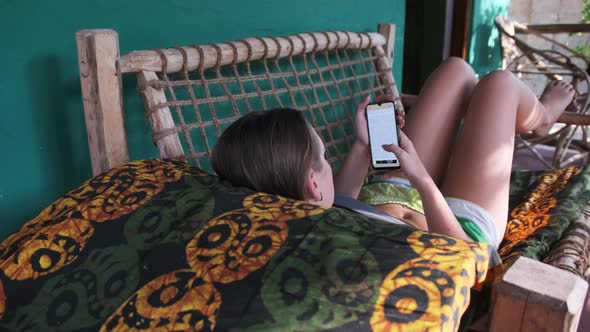 Young Girl Read Book From Smartphone Lying on Tropic Veranda of African Bungalow