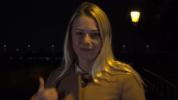 A Young Beautiful Woman Shows a Thumb Up To the Camera with a Smile and Nods in Urban Area at Night