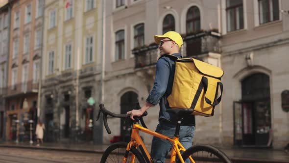 A Happy Delivery Man is Walking Through the Center of the Old Town and Holding a Bicycle with Him