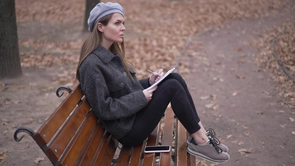 Thoughtful Inspired Woman Drawing Sketch Sitting on Bench in Overcast Autumn Park Looking Away