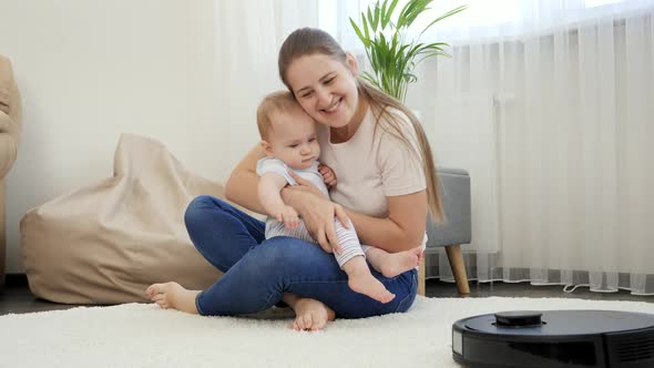 Smiling Mother and Baby Son Looking at Working Robot Vacuum Cleaner Cleaning Carpet in Living Room