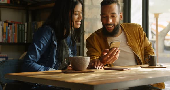 Front view of happy young mixed-race couple discussing over mobile phone at table in cafe 4k