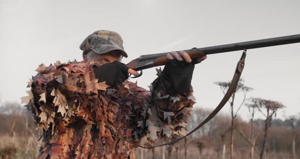 Portrait of Hunter in Hunting Equipment Aims With Shotgun Lies in Wait in the Field Sunset Light