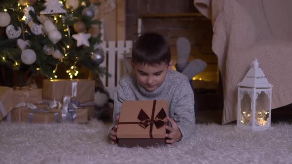 A Little Boy Opens a Box with a Gift and Rejoices Lying on the Floor Near the Christmas Tree