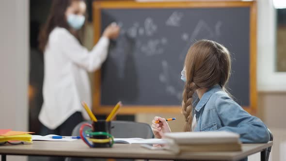 Portrait of Persistent Caucasian Schoolgirl in Covid Face Mask Writing in Workbook and Talking with