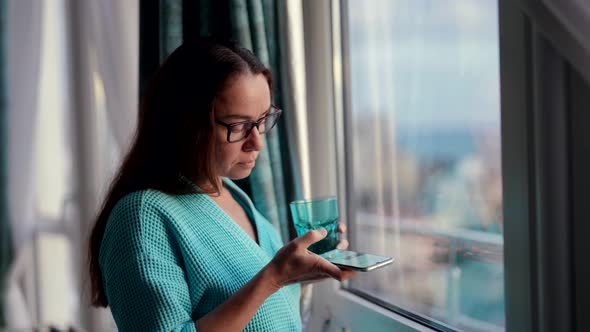 An Attractive Adult Woman with Glasses Standing at the Window with a Glass of Clean Water Holding a