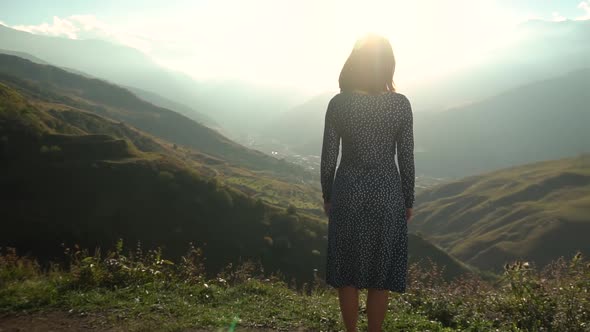 A Young Woman Stands in a Dress and Raises Her Hands Up Looking at the Mountains. The Sun Shines