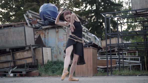 Wide Shot of Slim Elegant Ballerina in Black Dress Bound with Rope Dancing on Urban Slum