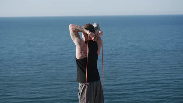 Backside View of Muscular Tattooed Sportsman Trains with Elastic Bands Outdoor