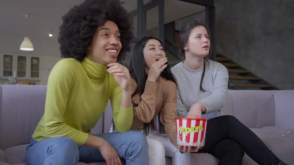 Three Cheerful Absorbed Multiethnic Women Watching Film Movie on TV Eating Popcorn and Talking in