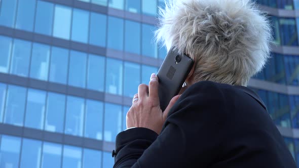 A Middle-aged Businesswoman Talks on a Smartphone in an Urban Area - a Windowed Office Building