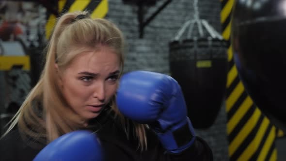 A Beautiful Young Woman in Blue Boxing Gloves Hits a Punching Bag in the Gym
