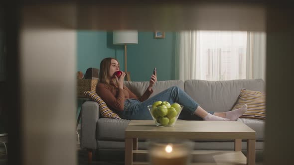 Young Woman Eats a Red Apple Lying on Sofa and and Speaks Via Video Link