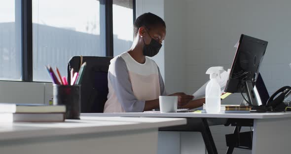 African american businesswoman wearing face mask, sitting at desk at work