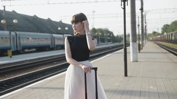 Glamorous Model in Blackwhite Dress Poses with Red Suitcase on Railway Platform