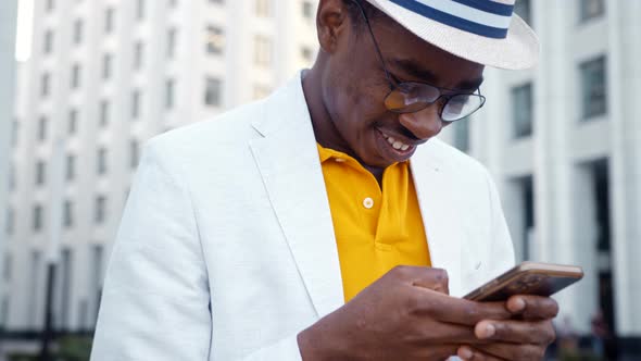 Joyful black man in glasses and suit types on smartphone standing