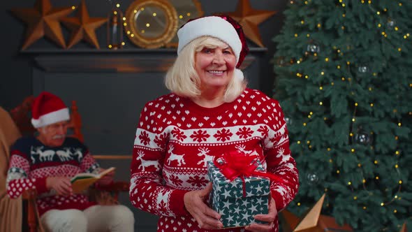 Mature Grandmother in Festive Sweater Presenting Christmas Gift Box Smiling Looking at Camera