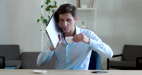 Young Funny Strange Business Man Joker Sits at Table, Drinks Coffee, Holds Laptop Near His Ear