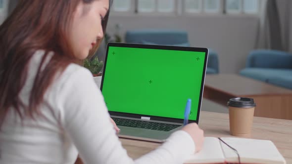 Girl Uses Laptop With Green Mock-Up Screen While Writing On Notebook In Her Living Room