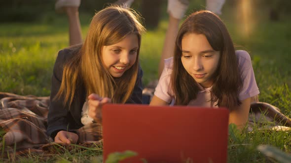 Two Adolescent Caucasian Girls Lying on Green Park Lawn in Sunbeam Talking Surfing Social Media on