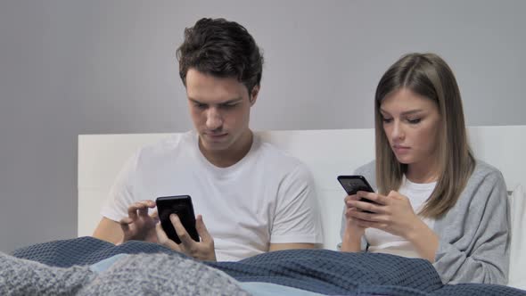 Young Couple Using Smartphone While Relaxing in Bed
