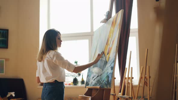 Busy Young Woman Is Casual Clothing Is Painting with Oil Paints Standing Near Easel and Holding