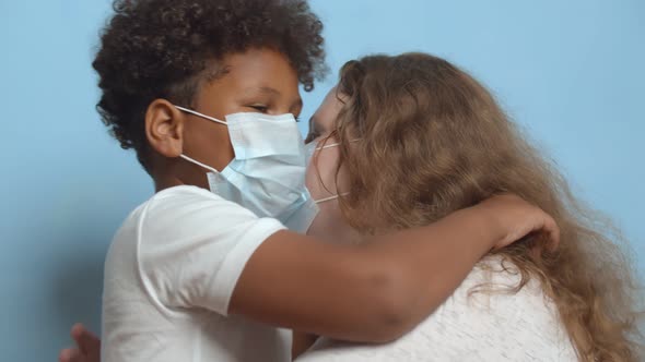 Side View of Little African Boy in Medical Mask Hugging Caucasian Mother Isolated on Blue Background