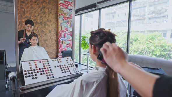 Asian stylist man using curling iron creating curly to young woman customer's hair in beauty salon.