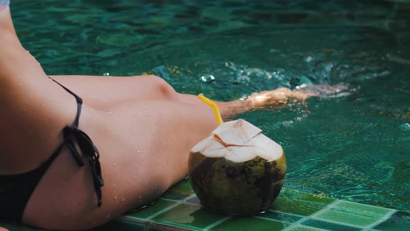 Closeup Legs of Tanned Young Woman Relaxing and Having Good Time Sitting on Poolside with Coconut