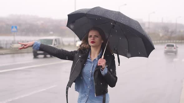 Beautiful Girl on the Road in Heavy Rain Under an Umbrella Trying To Catch a Car