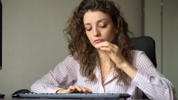 Young Female Office Manager in White Shirt and Curly Hair is Sitting at the Table and Typing Using