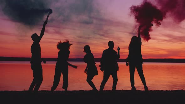 Joyful Friends Dancing Near the Lake at Sunset Holding Smoke Bombs Partying
