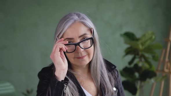 Confident Gray Haired Woman in Stylish Jacket Posing on Camera while Putting on Her Eyeglasses