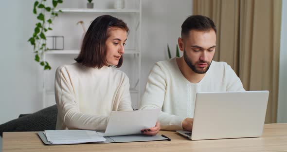 Serious Caucasiam Millennial Couple Sitting at Home Table Talking Holding Papers Discuss Pay