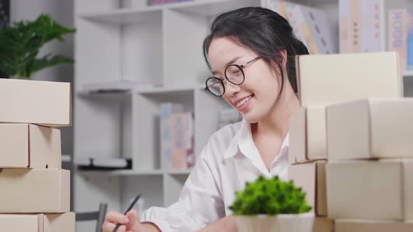 Happy young Asian woman wearing glasses, sitting and checking stock and taking a note