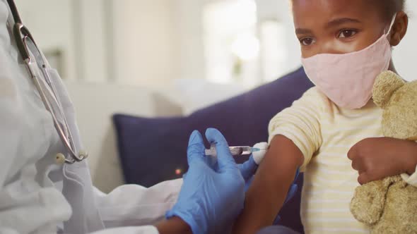 African american female doctor giving covid 19 vaccination to wearing face mask girl at home