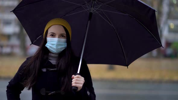 A Young Woman in a Protective Mask Walking in the Park Under Umbrella. Rainy Day, During Second Wave