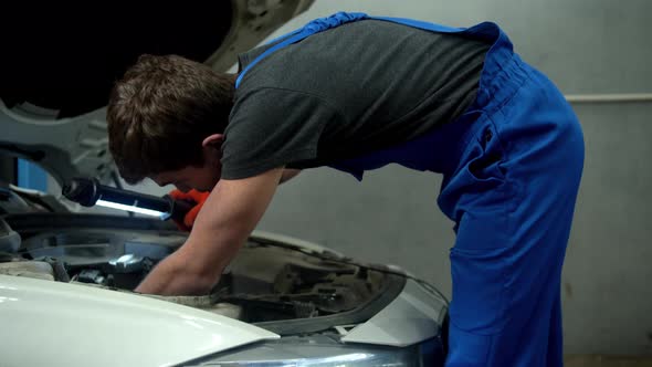 Mechanic with Flashlight Examines the Motor of Car