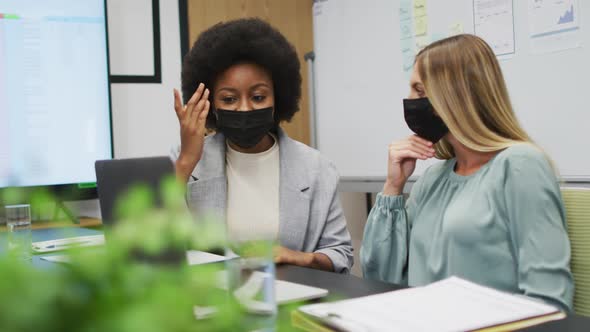 Two diverse businesswomen wearing face masks working together using laptop at desk in office