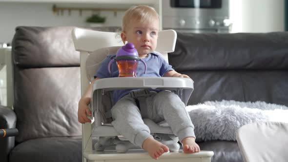 Cute Kid with Baby Straw Feeding Cup Sitting in Booster Seat One Year Old Toddler Watching Tv