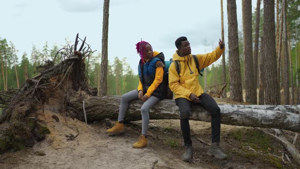 A Black Man and a Woman Sit on a Log During a Hike Relax and Take Selfies on a Mobile Phone