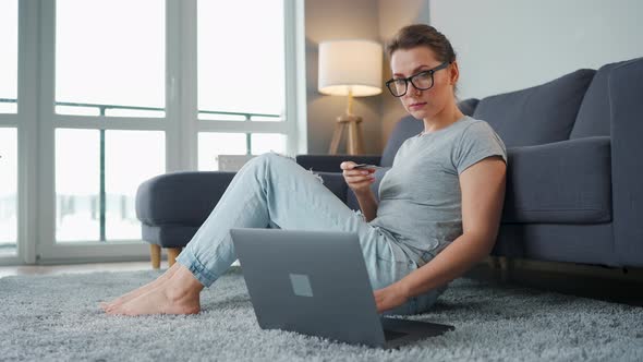 Woman with Glasses Sits on the Floor and Makes an Online Purchase Using a Credit Card and Laptop