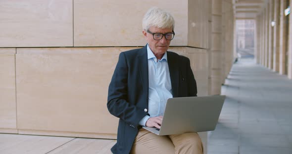 Portrait of Elegant Middle Aged Businessman Working on Laptop Sitting Near Modern Building Outdoors