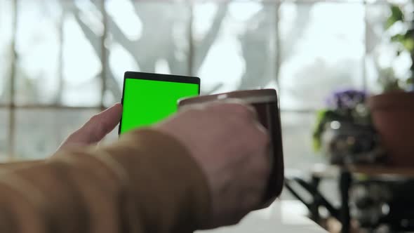 A Male Hand Holds a Phone with a Mockup Green Screen and Drinks Coffee